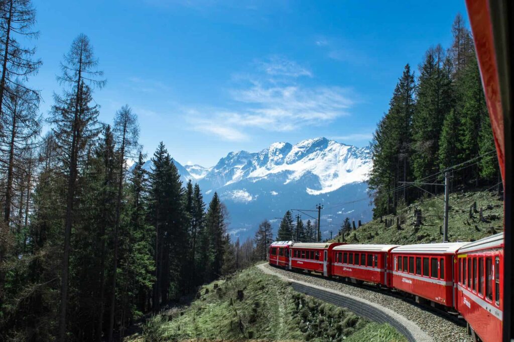 Red Bernina Express train crossing the iconic spiral stone viaduct with dramatic Alpine mountains and snow in background

